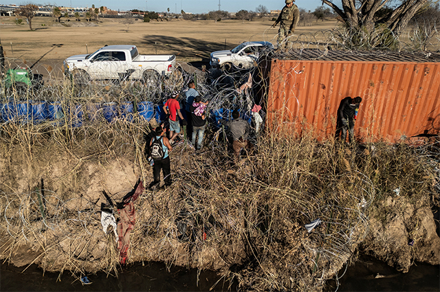 Солдат Национальной гвардии США стоит на транспортном контейнере, когда группа мигрантов пытается пройти через проволочное ограждение на берегу реки Рио-Гранде в Игл-Пасс, штат Техас.