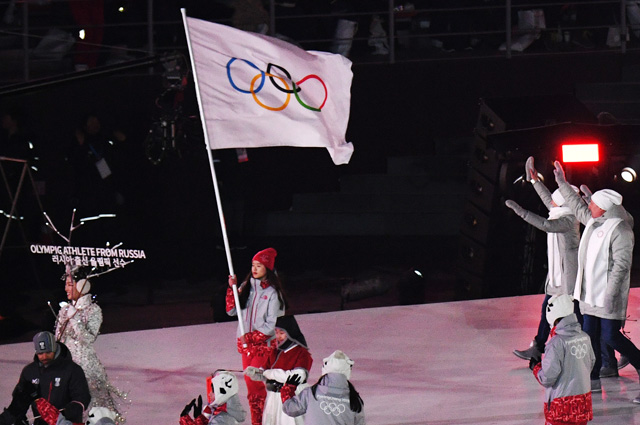 Волонтер несет олимпийский флаг и олимпийские атлеты из России, справа, приветствуют зрителей на церемонии открытия XXIII зимних Олимпийских игр в Пхенчхане.