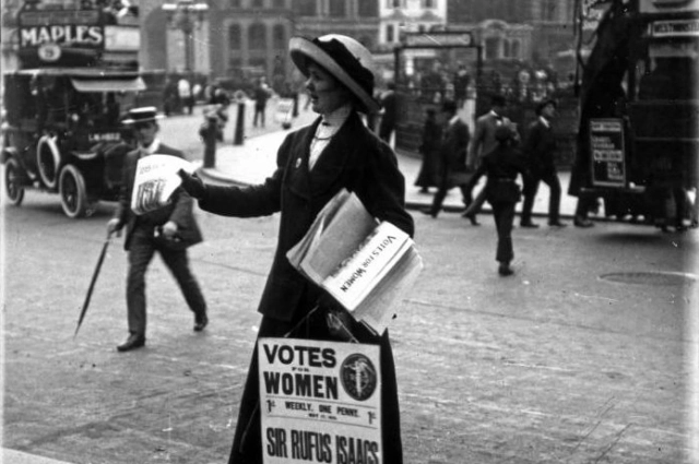 Британская суфражистка с плакатом раздает газеты или листовки на улице. Позади остановился двухъярусный автобус. Снято до 1919 года.